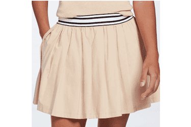 Model, shown from the waist down, wearing a beige Calia brand pleated tennis skirt and a matching top, hand in pocket of the skirt