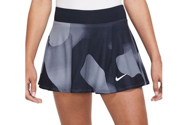 Model, shown from the waist down, wearing a dark blue and light blue printed Nike brand tennis skirt and a white top, hands at sides