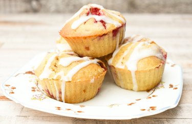 A closeup of three vegan raspberry muffins drizzles with white frosting on a floral plate.