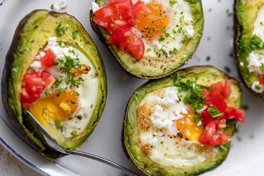 Baked eggs in avocados topped with dices tomatoes, herbs, salt and pepper on a white plate