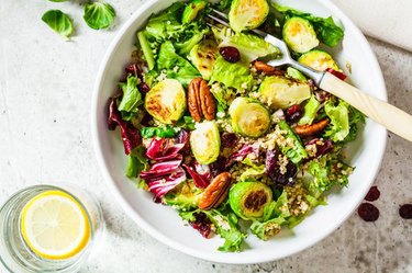Fried brussels sprouts salad with quinoa, cranberries and nuts in white bowl, top view.