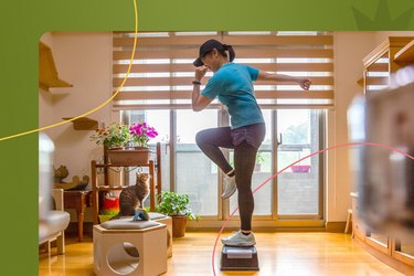 An athlete wearing a blue shirt, leggings and black hat is doing a step-up exercise at home while a tortoiseshell cat looks on during the 4-week legs challenge