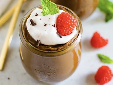 A glass jar of avocado chocolate pudding topped with whipped cream, raspberries and mint