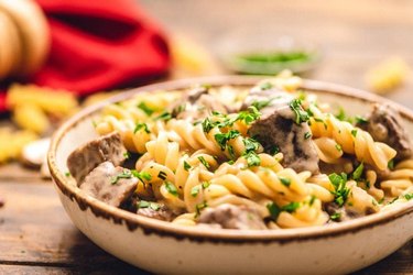 Egg noodles with beef pieces in brown bowl on wooden background.