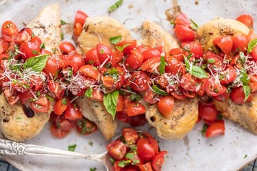 Chicken breast topped with baby tomatoes, chopped basil and parmesan cheese on a white plate.