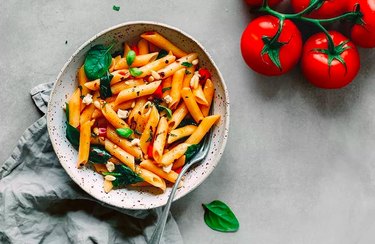 Penne pasta with spinach and diced tomatoes in a white speckled bowl on gray background.