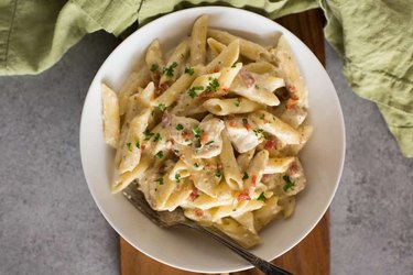 Penne pasta in ranch sauce with bacon and chives in a white bowl on cutting board background.