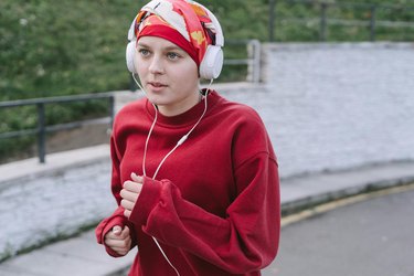 a young adult wearing a red sweatshirt, a red patterned headscarf and white headphones jogs outside