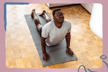 A young Black man who is wearing glasses and grey workout clothes closes his eyes and holds cobra pose on a dark grey yoga mat as part of the 30-Day Yoga Challenge