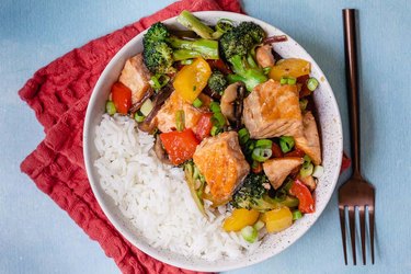 Stir Fry Salmon with vegetables on top of white rice on a white plate with a fork