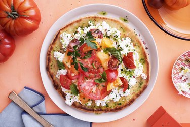 overhead photo of round tomato and cheese flatbread on white plate on peach table with tomatoes and cloth napkins