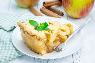 A slice of apple pie on a table with fresh apples and cinnamon.