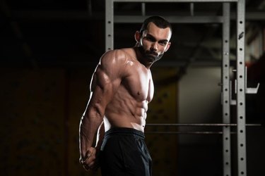 Youn, muscular man with a beard standing in a dark gym, flexing his biceps, shoulders, triceps, pecs and abs.