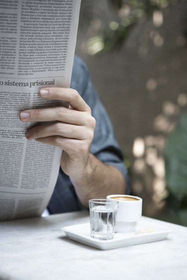 Man reading newspaper outdoors (focus on cups of espresso and water)