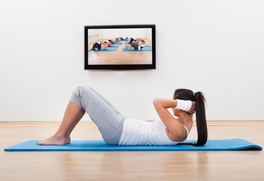 Athletic barefoot young woman working out at home lying on a mat doing liftups and head raises while watching and participating in a class