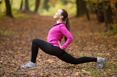 Sporty beautiful happy young woman practicing yoga, standing in low lunge exercise, anjaneyasana, ashwa sanchalanasana, working out outdoor on autumn day wearing sportswear sweatshirt. Full length