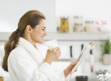 Happy housewife in bathrobe drinking milk and reading magazine