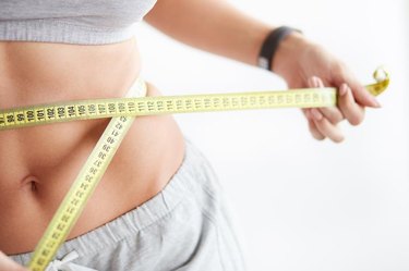 Bright closeup of slim young woman measuring her thin waist with a tape measure on white background