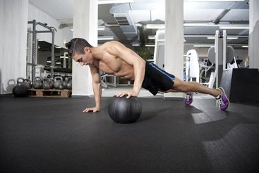 Fit and attractive athlete is performing a one-handed push-up in a medicine ball.