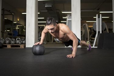 Fit and attractive athlete is performing a one-handed push-up on a medicine ball at the gym.