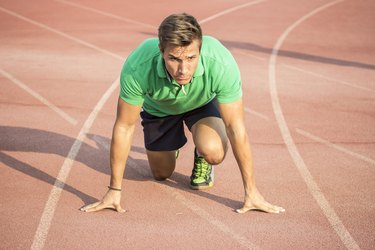 Male sprinter in clothes at start position, on red track.