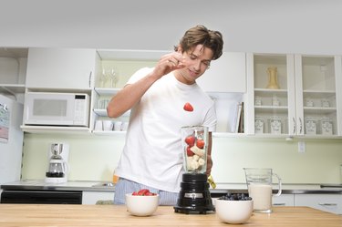 Young man in kitchen, dropping strawberry into fruit filled blender