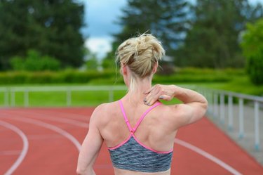 Rear view of athletic female sprinter massaging her own neck at race track with trees in background