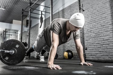 Nice guy with a beard prepares to make a pushup in the gym on the gray brick wall background. He wears sportswear, white sneakers and a white cap. Man looks at the floor. Horizontal.