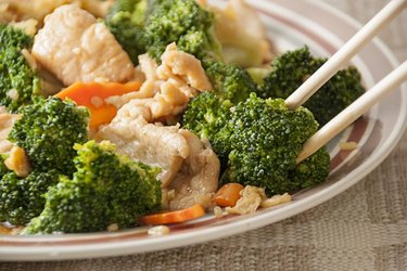 Closeup of an individual serving of chicken and broccoli, with very selective focus on the broccoli floret that is being held by the chopstick