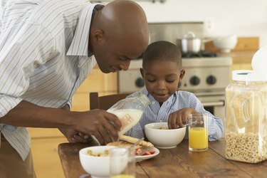 Father pouring milk in bowl for son (5-7) at breakfast table