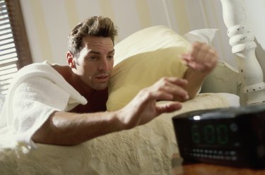 Young man lying on a bed reaching for the alarm clock