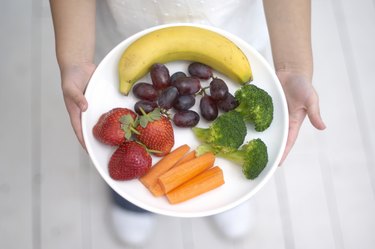 Girl holding plate of fruit and vegetables, including banana, strawberries, grapes, broccoli, carrots