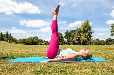 gym and pilates outside - active young blond woman exercising, lying down with both legs up and arms down to tone them up on her exercise mat over sunny blue sky