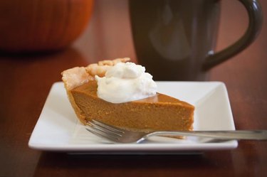A slice of pumpkin pie with a daub of whipped cream on a white square plate with a fork and coffee cup.
