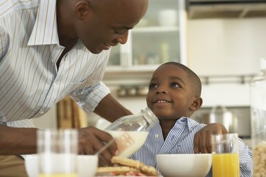 Father pouring milk in bowl for son (5-7) at breakfast table, smiling