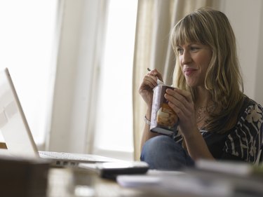 Young woman looking at laptop, holding ice cream carton, smiling