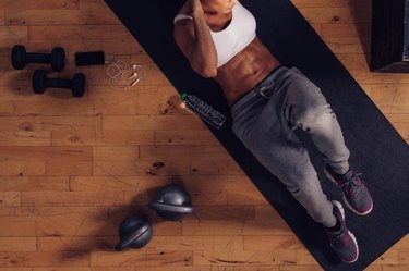 Top view of muscular woman doing sit-ups. Female lying on exercise mat doing stomach workout with gym equipments in floor.