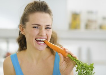 Happy young woman eating carrot in kitchen