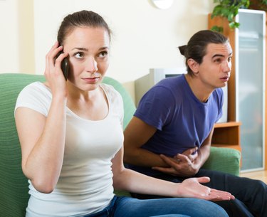 A woman talks into a smart phone as her boyfriend clutches his tummy on the couch.