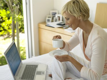 Woman sitting on bed holding cup and saucer, looking at laptop