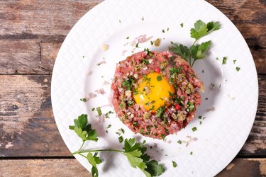 steak tartare topped with a raw egg spices and parsley on a white plate over wooden background.