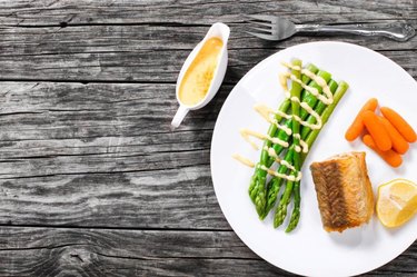 Hake served with asparagus, piece of lemon, baby carrots and hollandaise sauce on a white platter atop an old rustic table