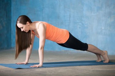 Beautiful young woman dressed in bright sportswear doing core exercise indoors. Yogi girl working out in grunge interior with blue wall. Standing in plank pose. Full length. Side view
