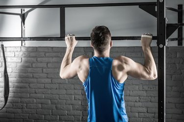 Active man makes a pull-up on the crossbar in the gym on the gray brick wall background. He wears a blue sleeveless. Shoot from the back. Horizontal.