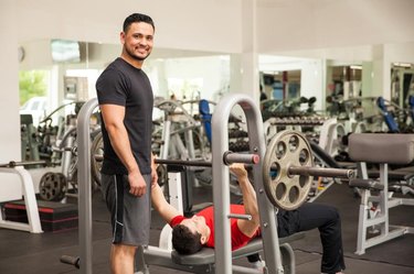 Portrait of a handsome young Hispanic man spotting his friend on the bench press while working out at a gym