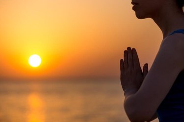 Profile of serene young woman relaxing on the beach, meditating with hands in Namaste gesture at sunset or sunrise, close up