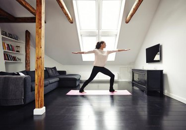 Full length image of fit woman standing on exercise mat with arms outstretched doing yoga in loving room. Caucasian female model exercising at home.