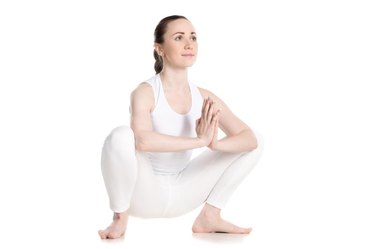 Sporty smiling beautiful young woman in white sportswear sitting in squat, Garland Pose, Malasana with hands in Namaste, studio full length shot on white background, three-quarters view, isolated