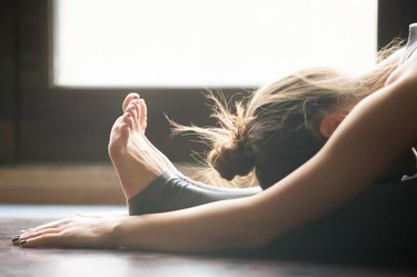 Young woman practicing yoga, sitting in Seated forward bend exercise, paschimottanasana pose, working out, wearing sportswear, grey pants, bra, indoor, home interior background, close up