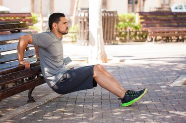 Profile view of a strong man exercising his arms and doing tricep dips outdoors in a park bench